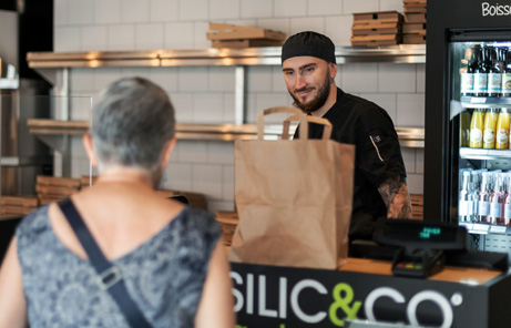 Photo d'Alexis Segura, franchisé Basilic & Co derrière son comptoir. Il sert une cliente que l'on voit dos au comptoir.